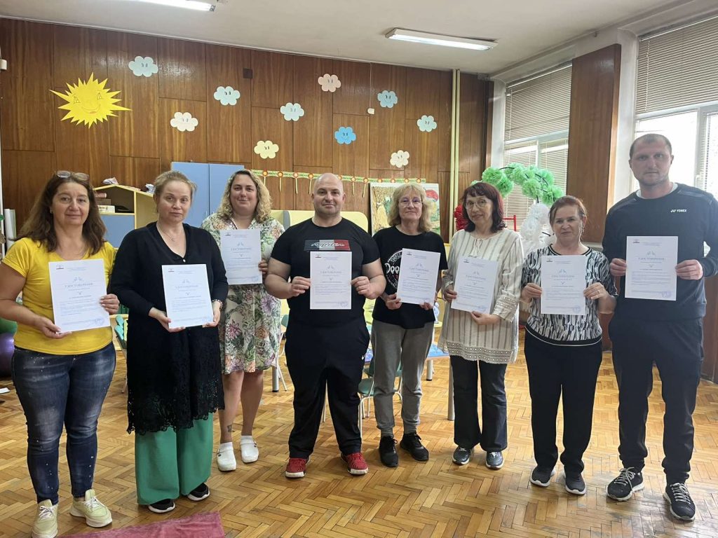 Eight adults stand in a classroom, each holding a certificate and smiling, with sun and cloud decorations on a wood-paneled wall behind them.