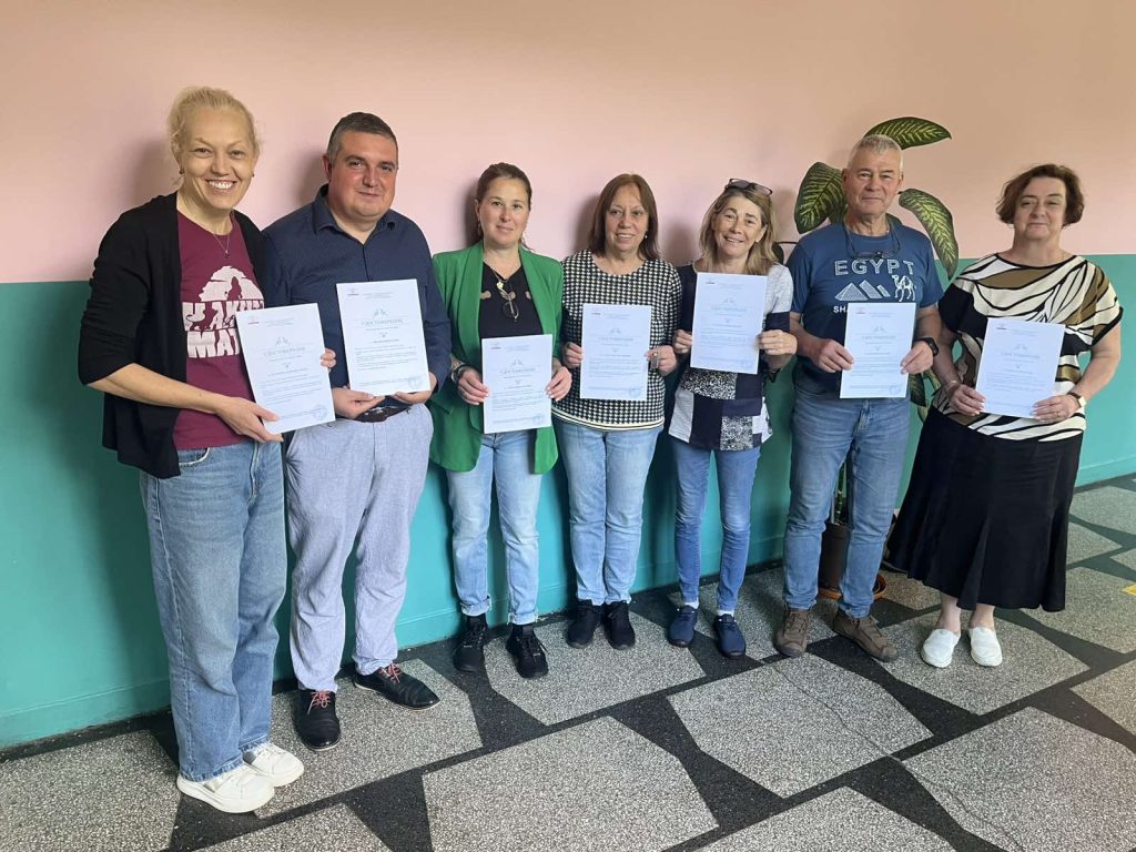 Seven adults stand in a hallway, each holding a certificate and smiling at the camera.