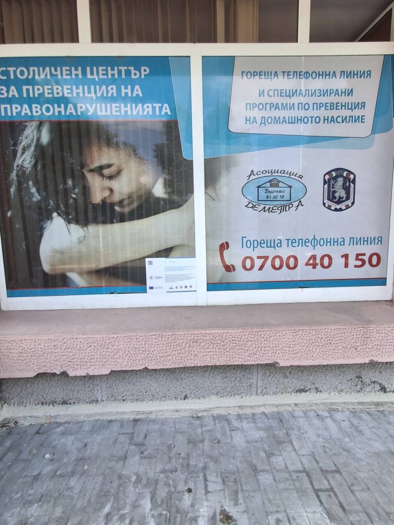 Poster on a storefront window advertising domestic violence prevention services, including a helpline number 0700 40 150 and organizational logos.